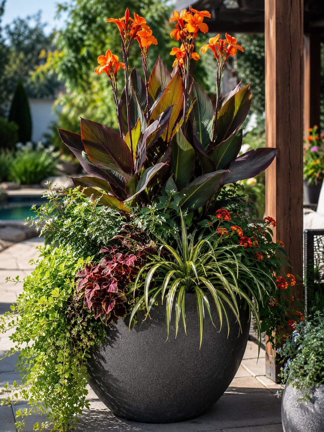 A large dark gray stone planter filled with a mixed garden arrangement, including canna lilies with orange flowers, dark burgundy foliage, green trailing plants, and a spiky green-and-white center plant, set outdoors beside a wooden post and pool area.